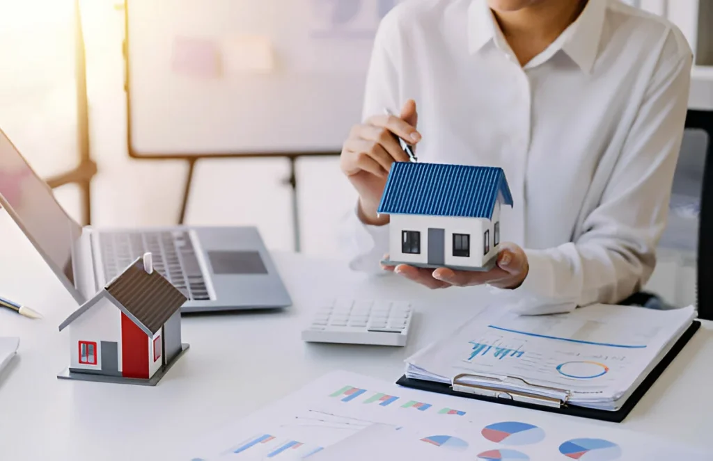 Person holding house model with charts, laptop, and documents on desk.