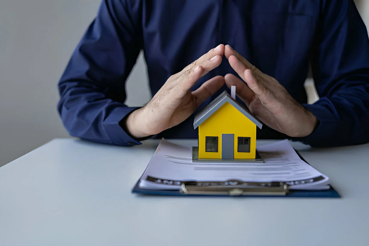 Hands shielding a yellow house model placed on documents.