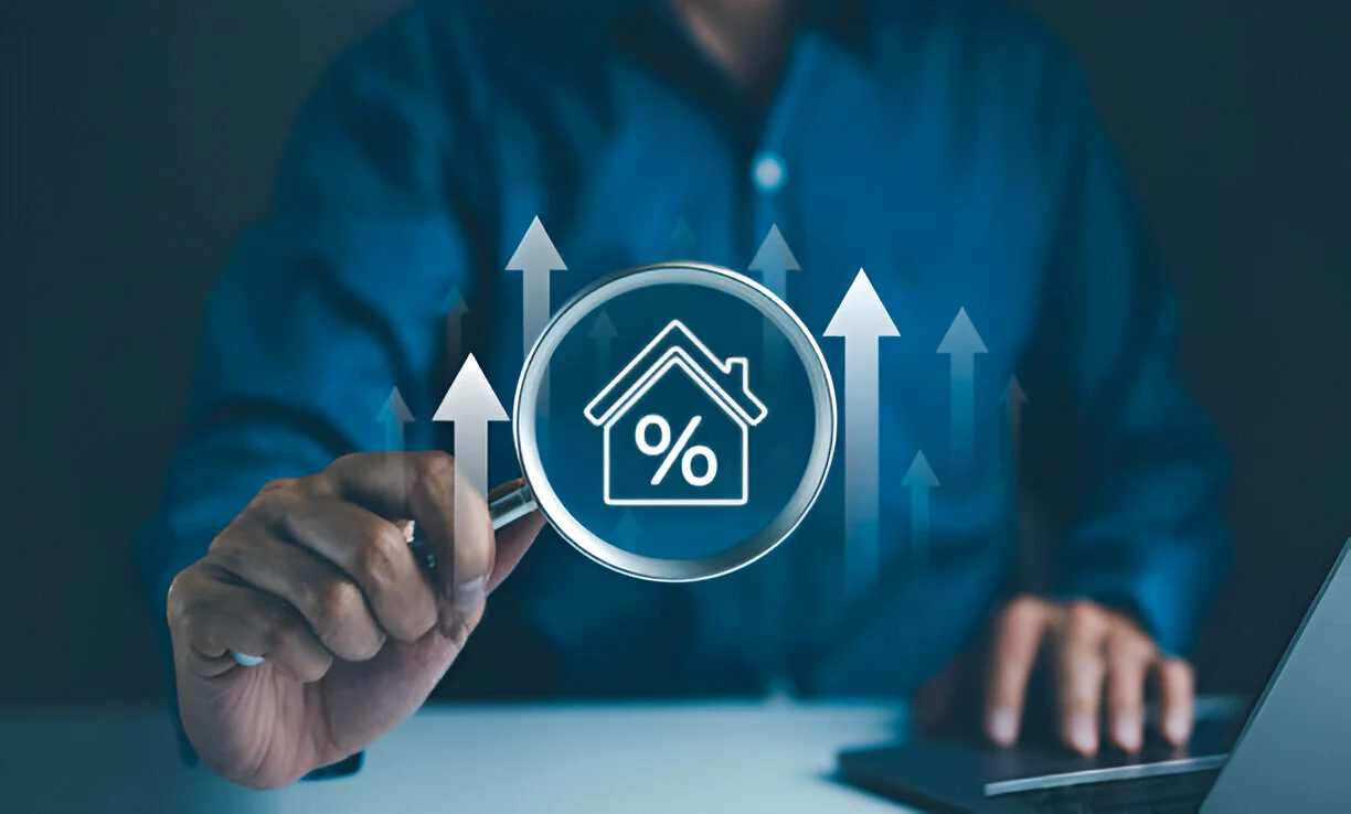 Person using magnifying glass to examine house interest rates with upward trend arrows.